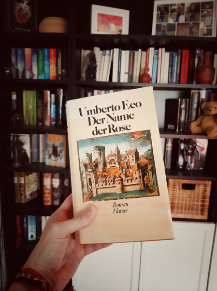 Eine Hand vor einem Bücherregal hält ein Buch in die Kamera: "Der Name der Rose" von Umberto Eco, erschienen im Carl Hanser Verlag.