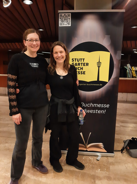 Lektorin und Autorin Harlow Alexander (links) mit Sonja Tornefeld (rechts) stehend vor dem Roll-Up der Stuttgarter Buchmesse