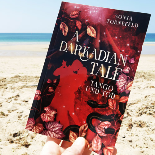 Eine Hand hält das Buch "A Darkadian Tale Tango und Tod" vor einem Strandhintergrund in die Kamera. Das Cover ist in Rottönen gehalten. Es zeigt links ein tanzendes Paar als Silhouette, umrahmt von rotgoldenen Weinblättern. Rechts unten sieht man grafisch eine schwarze Schlange, im Hintergrund ein Zirkuszelt, das im Hintergrund verschwindet.