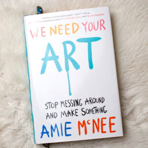 Buch "We need your art" von Amie McNee liegend auf weißem Fell fotografiert. Das Cover ist weiß, die Schrift in bunten Buchstaben, die wie selbstgemalt aussehen. Vom R in Art fließt Farbe herunter.