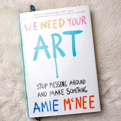 Buch "We need your art" von Amie McNee liegend auf weißem Fell fotografiert. Das Cover ist weiß, die Schrift in bunten Buchstaben, die wie selbstgemalt aussehen. Vom R in Art fließt Farbe herunter.